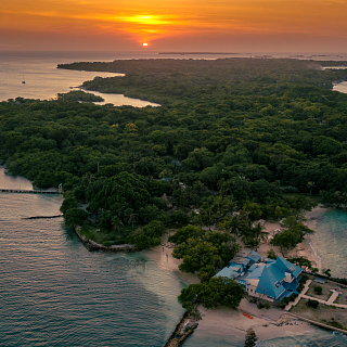Espectacular Atardecer sobre Isla Tropical en el Caribe colombiano Vista aérea panorámica de isla tropical cubierta de vegetación al atardecer, con un edificio de techo azul en la punta, muelles y el sol poniéndose sobre el océano tranquilo.