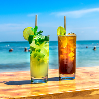 Refrescantes Cócteles Tropicales en Barra de Playa Paradisíaca Refrescante mojito y cóctel oscuro tipo té helado en vasos altos sobre barra de madera, con fondo de playa tropical de agua turquesa y cielo azul
