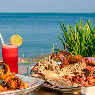 Parrillada de Mariscos Frescos con Langosta Frente al Mar Deliciosa parrillada de mariscos frescos con langosta y mejillones, servida con bebidas rojas en una mesa de madera con vista al mar azul